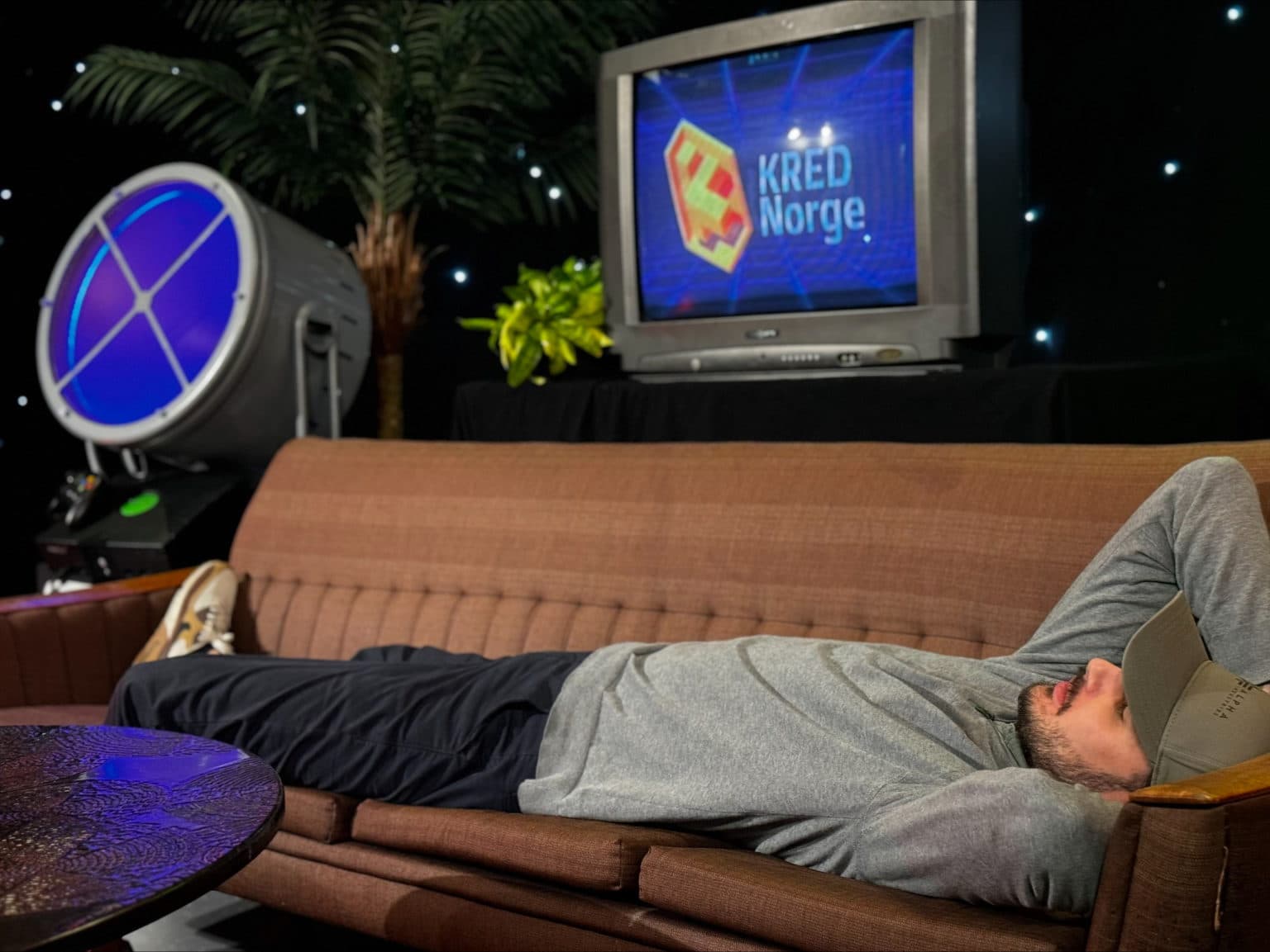 Person ligger avslappet på brun sofa med hendene bak hodet, iført grå hettegenser og caps. I bakgrunnen vises spotlight med blå linse, palmeplante og TV-skjerm med teksten "KRED Norge" og fargerik logo.