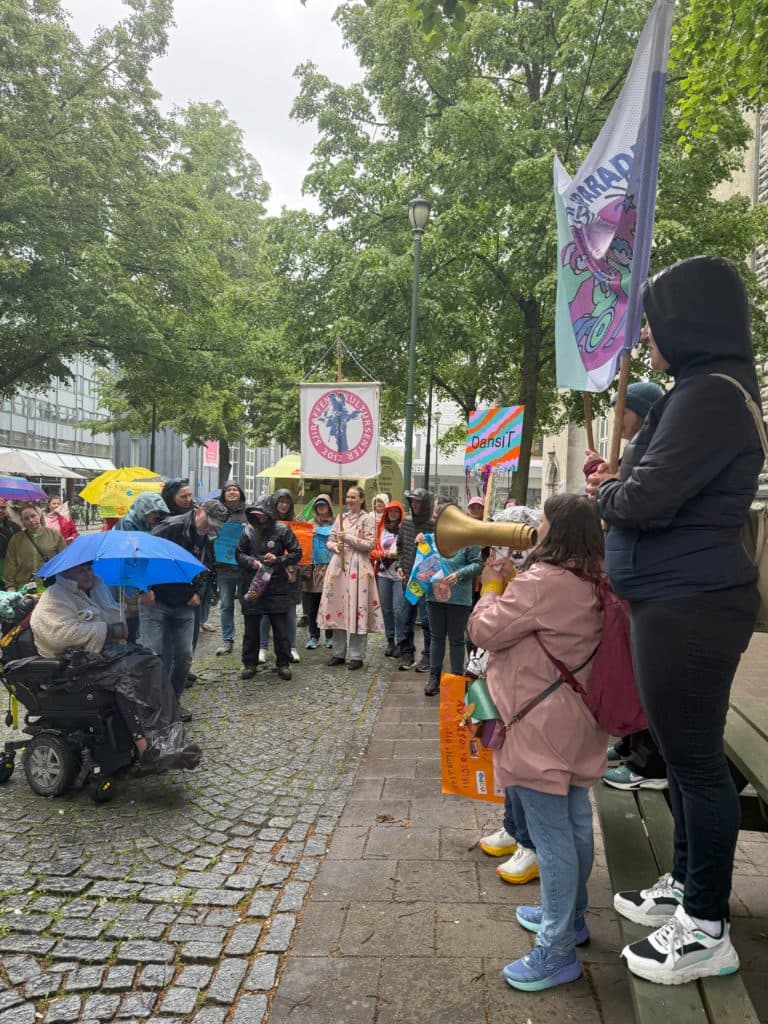 Person står på benk og snakker i megafon under en demonstrasjon på brosteinsgate omgitt av trær. Ved siden av holdes et flagg med tegneseriepreg og teksten "PARADE".  En person i elektrisk rullestol holder blå paraply.