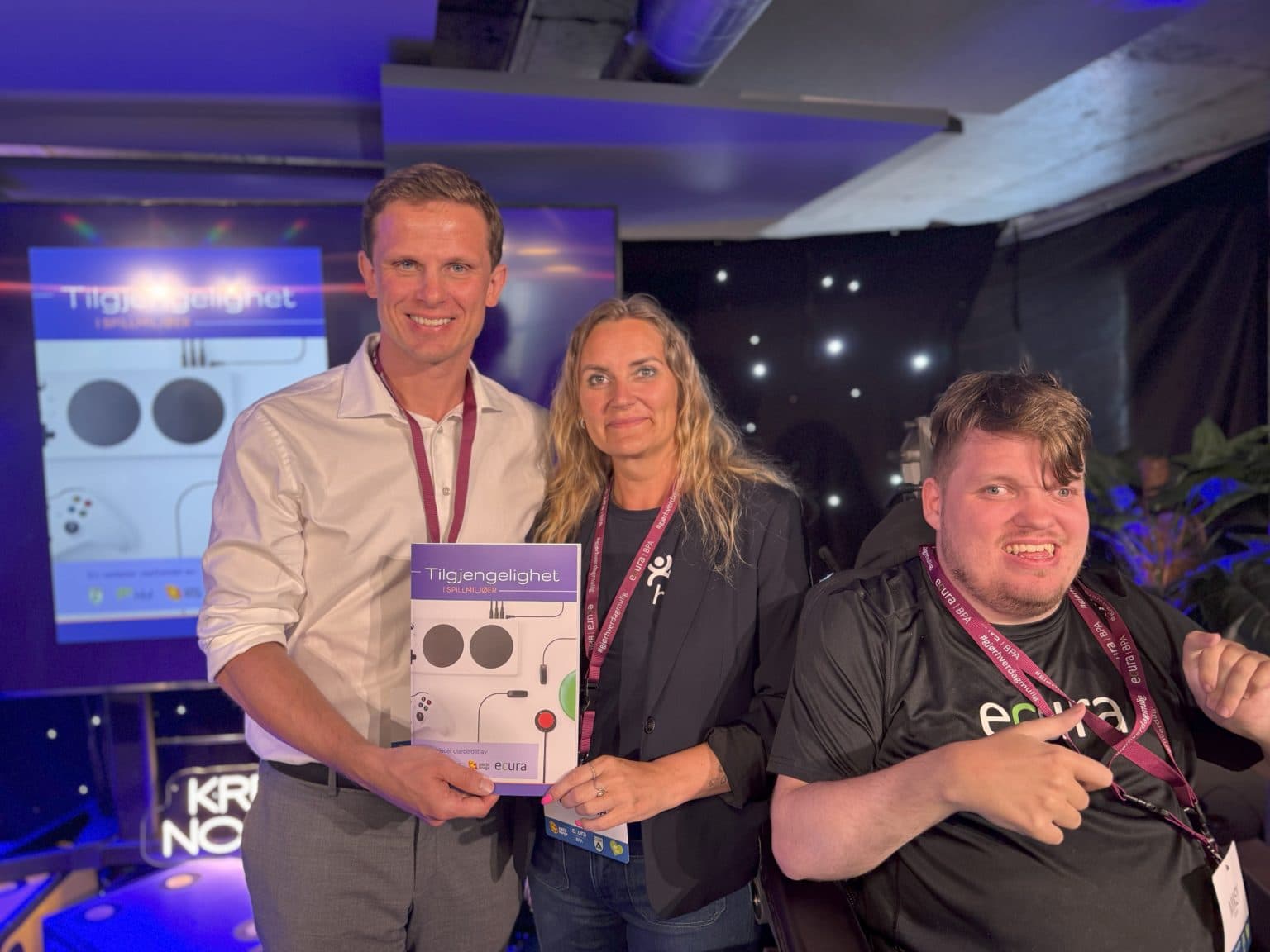 Tre personer poserer med veilederen «Tilgjengelighet i spillmiljøer», én i rullestol og én med brosjyren i hånden – et bilde på samarbeid og lansering av inkluderende tiltak.