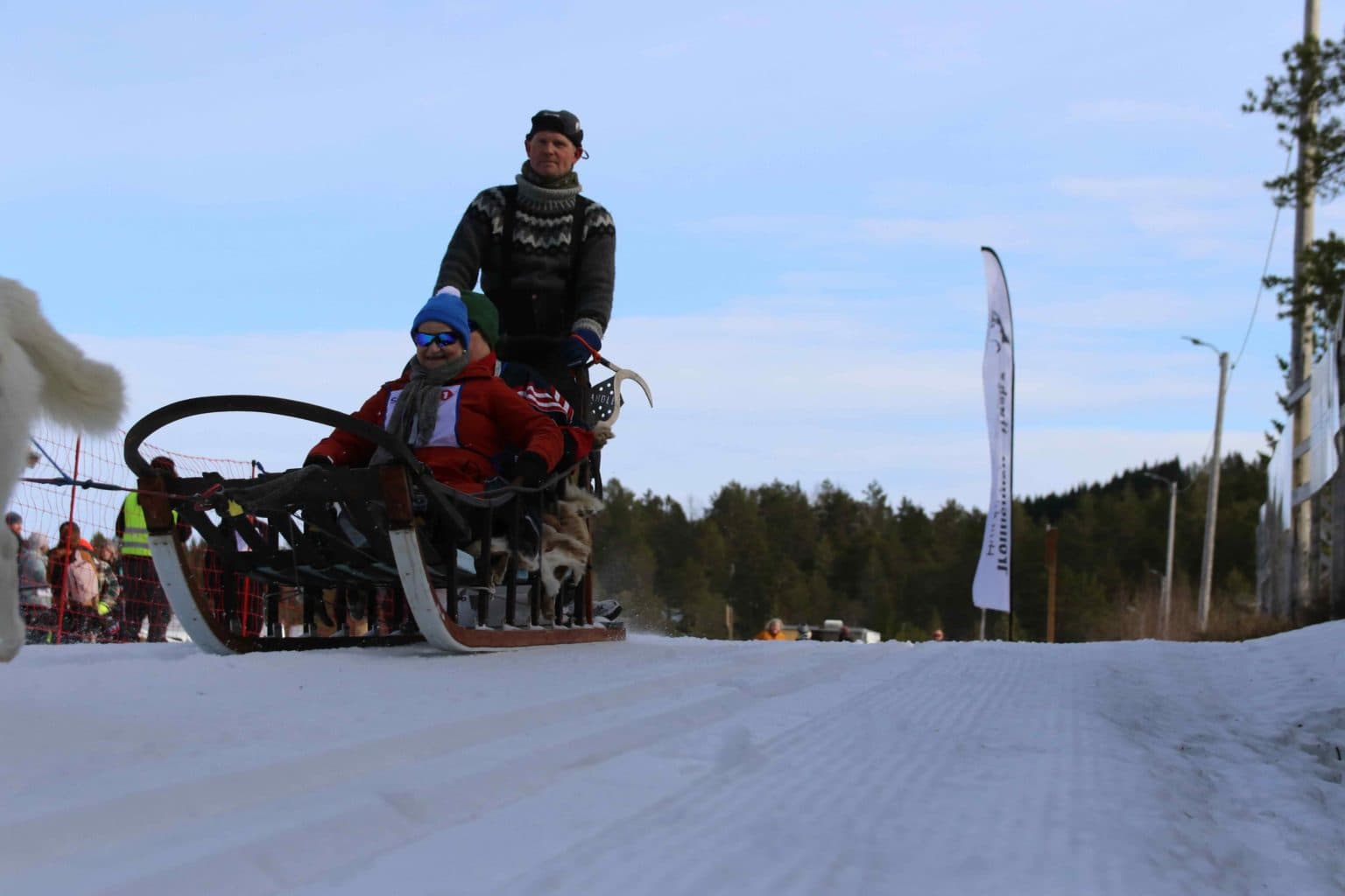 Tre personer kjører hundeslede på snødekt sti, med hunder delvis synlige foran. Én står bak som fører, to sitter foran med vinterklær og solbriller. Bak et sikkerhetsnett står publikum og banner.
