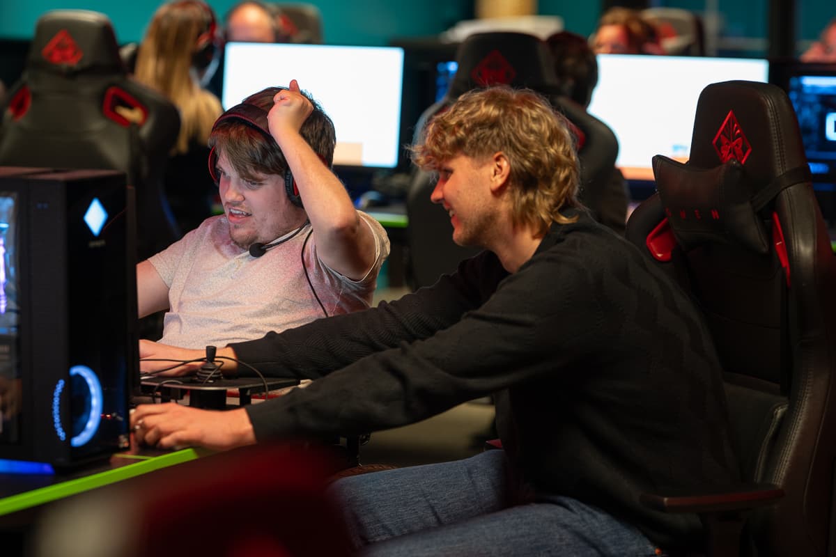 To personer i OMEN-gamingstoler deltar i dataspill med headset. Den ene reagerer intenst, den andre smiler. Bak dem sees flere spillere i lignende oppsett.