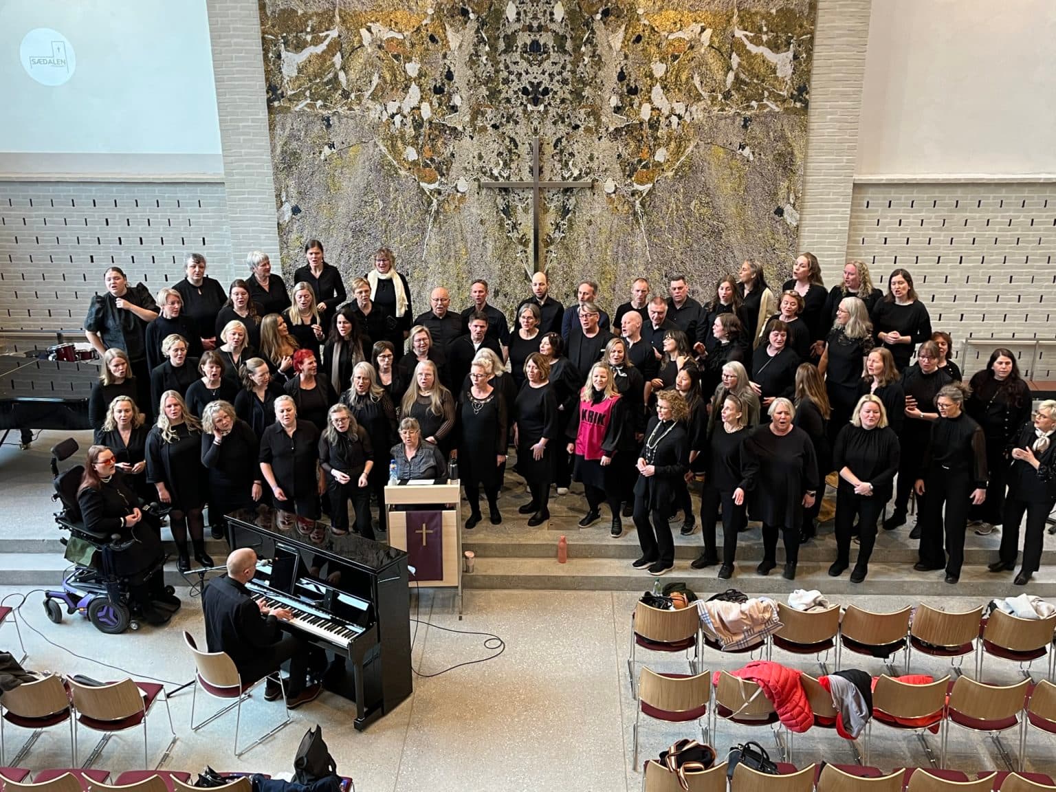 Stort kor i svarte klær står samlet foran et kors i en kirke. En pianist spiller ved et svart piano, og en kvinne ved en talerstol med lilla klede leder gruppen. Tomme stoler med jakker og vesker står i forgrunnen, og rommet har en høytidelig atmosfære.