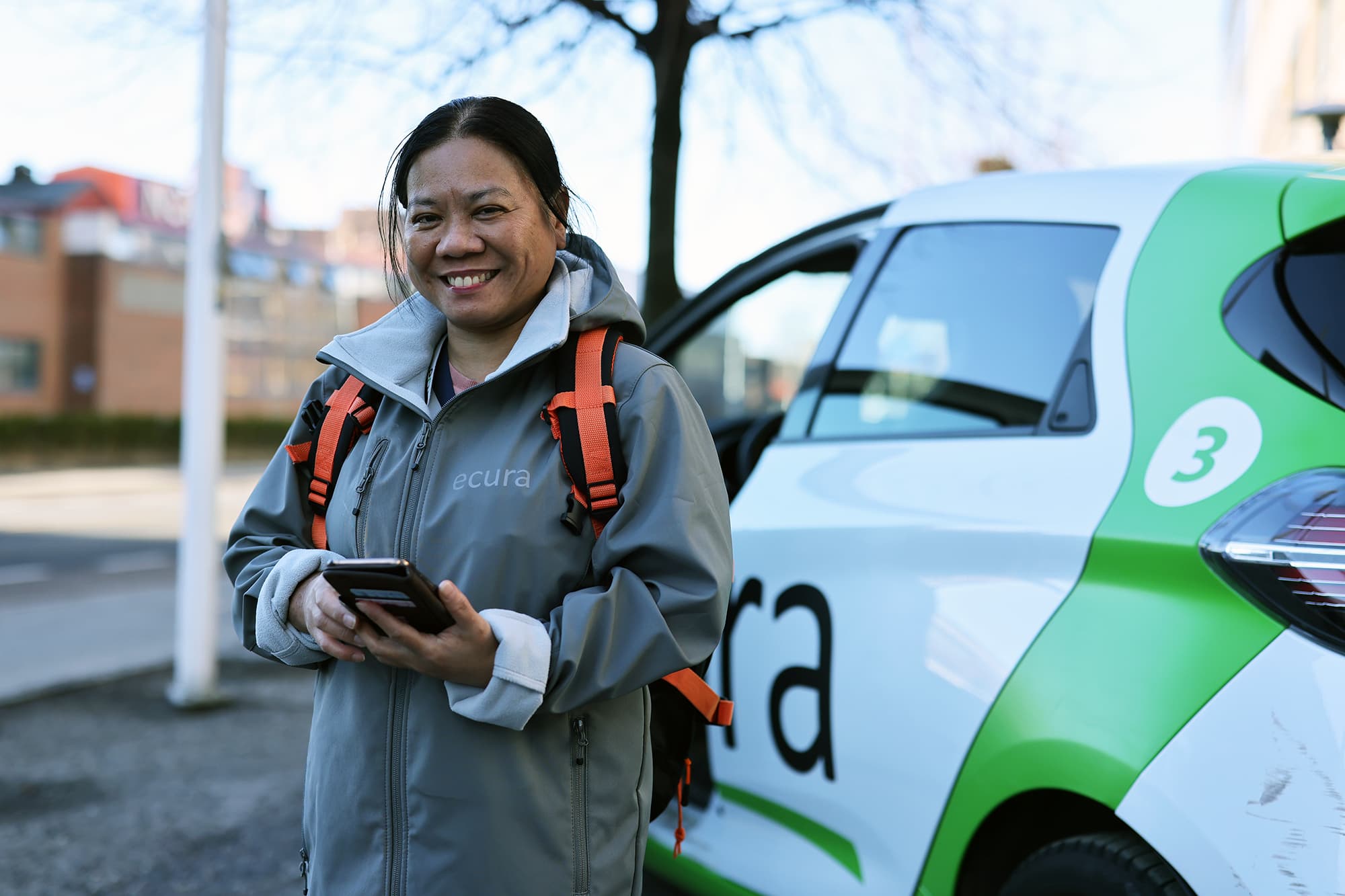 Smilende person med oransje ryggsekk og 'ecura'-jakke står ved en hvit og grønn firmabil med nummer 3, i urbant miljø med trær og bygninger i bakgrunnen.