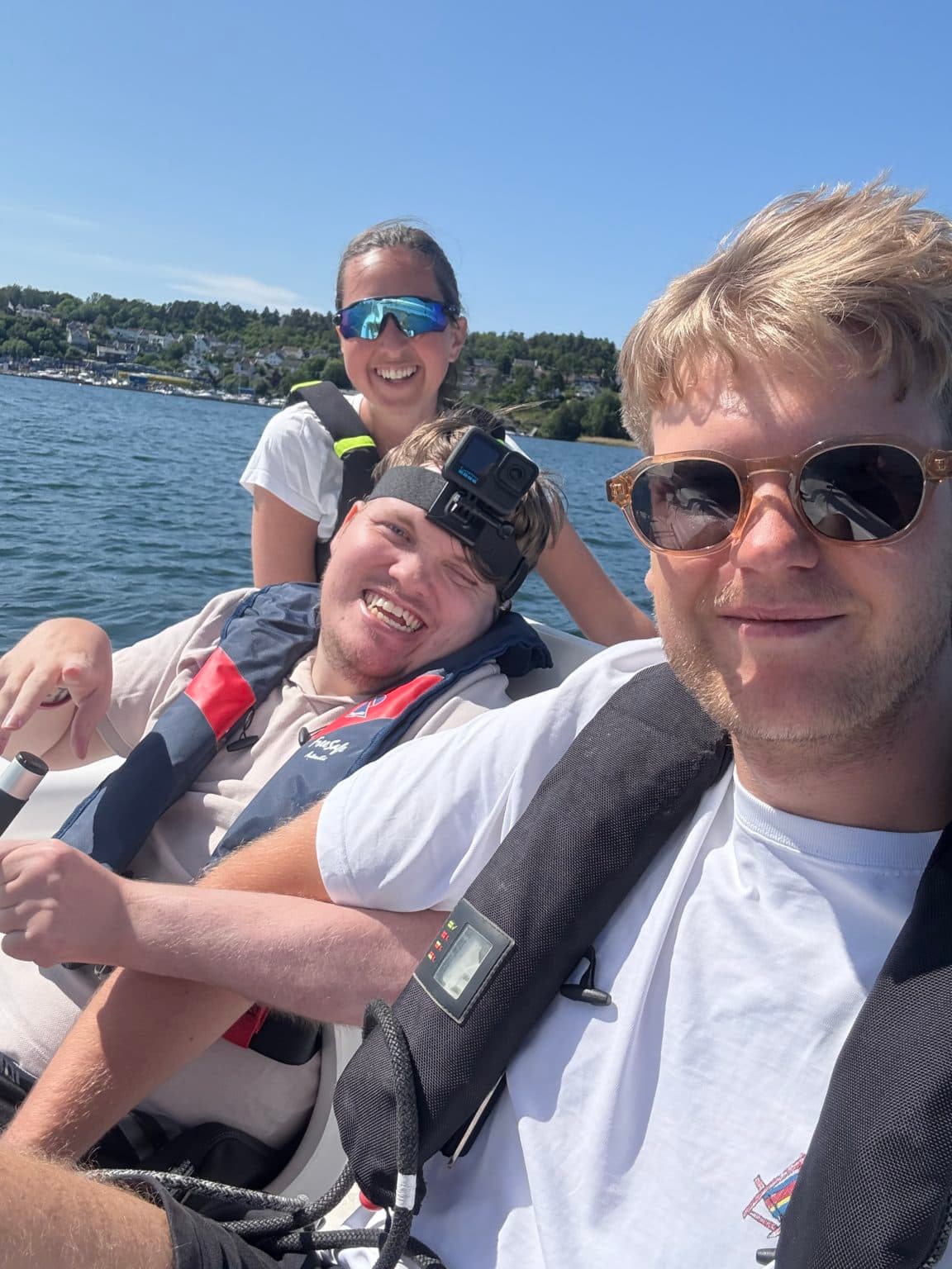 Tre personer med redningsvester nyter en solrik båttur. Én har GoPro-kamera festet på hodet. Bak dem sees kystlandskap med hus og trær, og vannet er rolig under blå himmel.