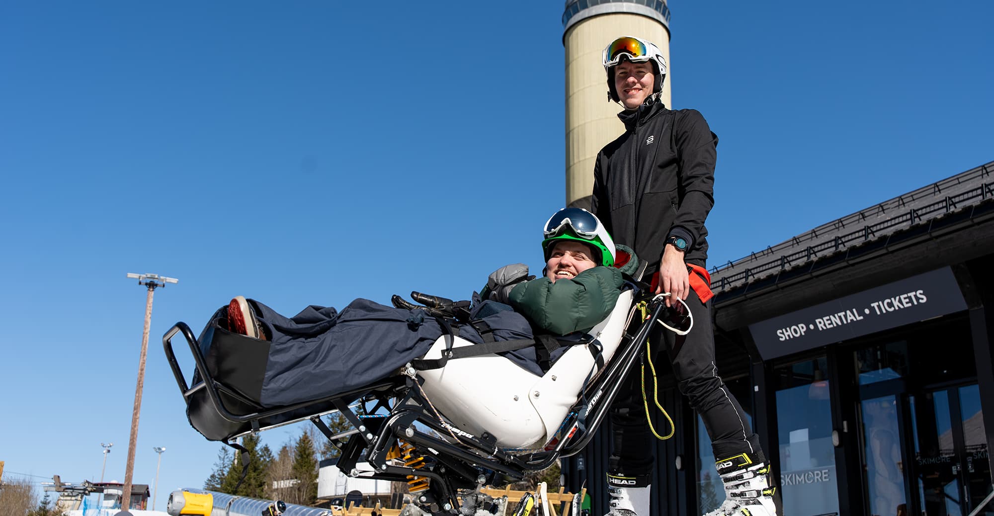 To personer ved skiutleie – én i sitski, én stående. Vinterklær og blå himmel.