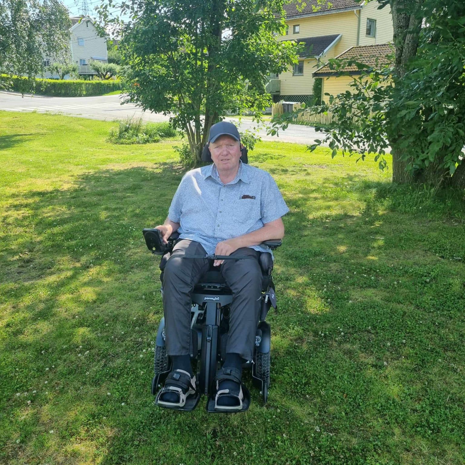 Person i motorisert rullestol på gressplen med trær og hus i bakgrunnen. Lys blå skjorte, mørke bukser og caps.