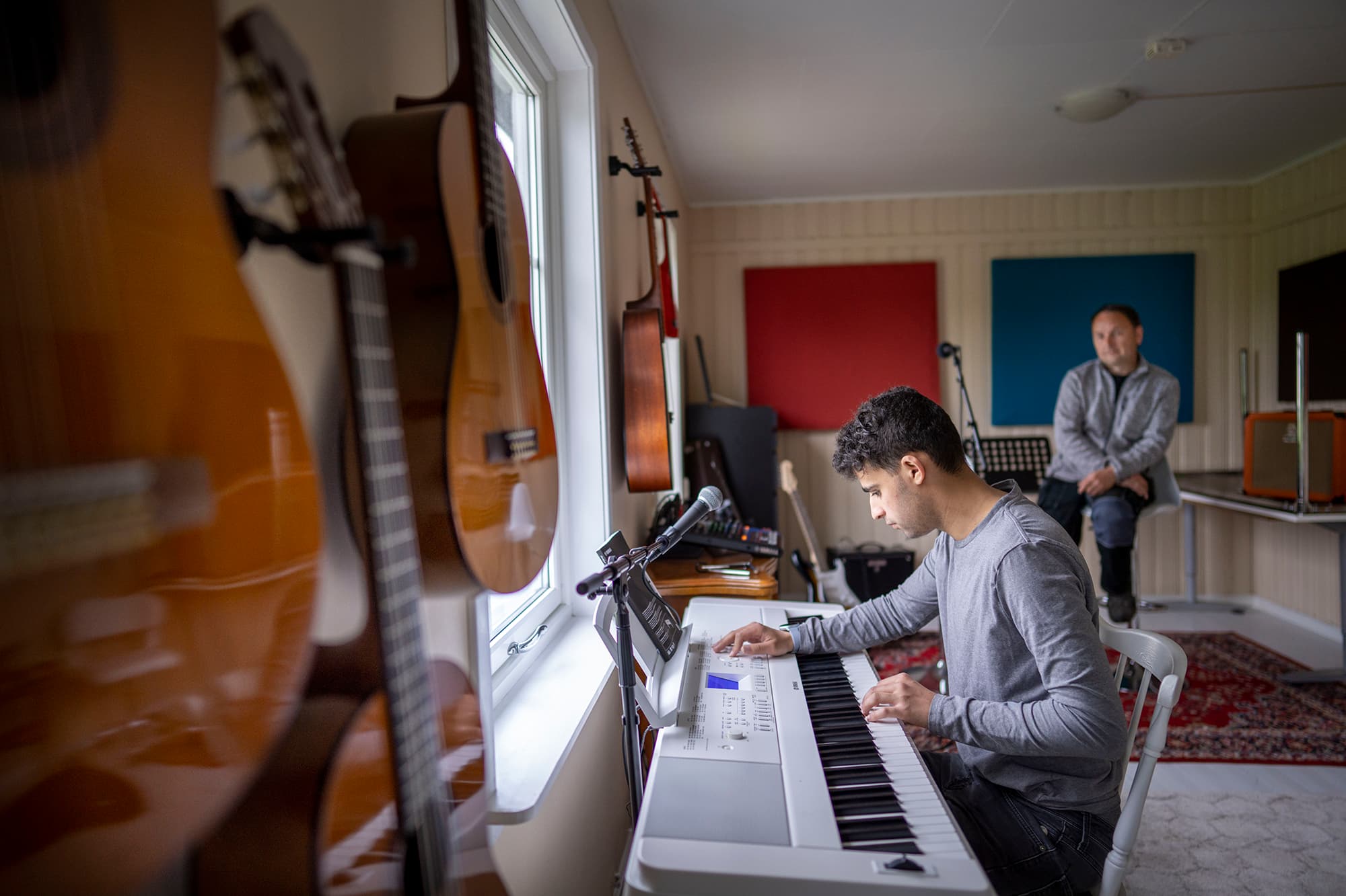 To personer i musikkrom med gitarer på veggen og digitalt keyboard. Én spiller, én lytter. Fargerike lydpaneler, mikrofonstativ og musikkutstyr skaper kreativ atmosfære.