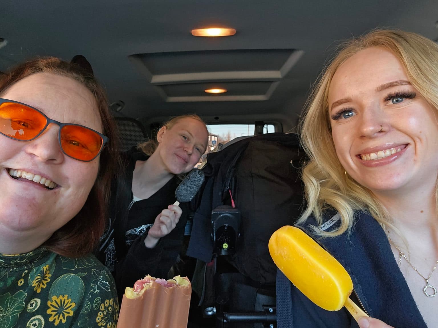 Tre personer smiler og holder ispinner inne i en bil. Den ene har oransje briller og blomstrete topp, den andre holder en sjokoladeis bak en stor bag, og den tredje har langt blondt hår og mørk jakke.
