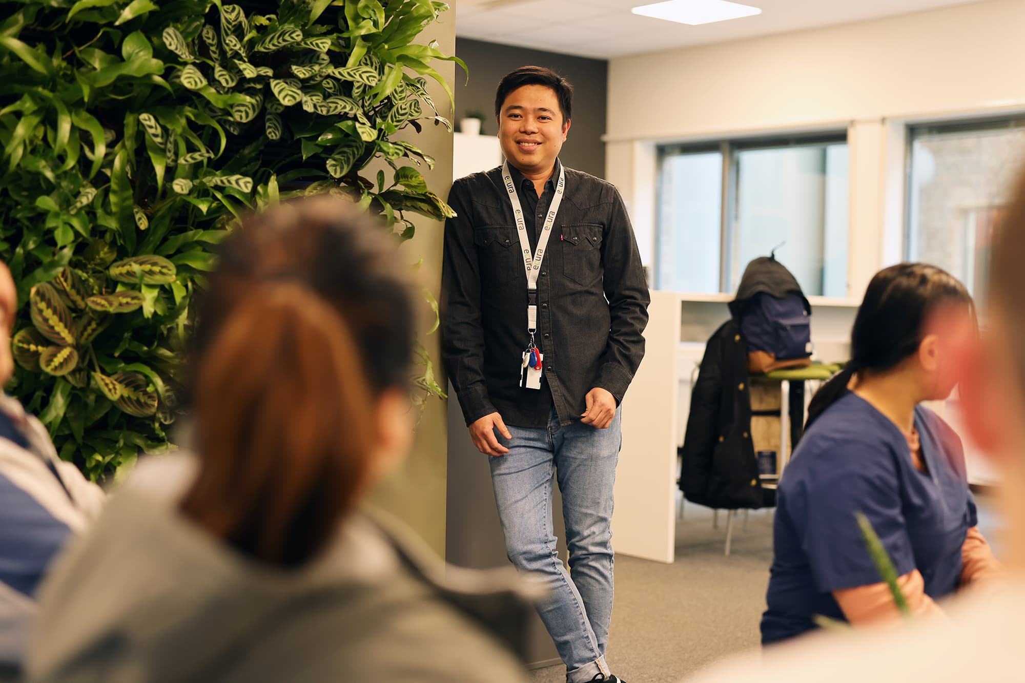 Person i svart skjorte og lys jeans lener seg mot vegg, omgitt av grønn innredning og kolleger.