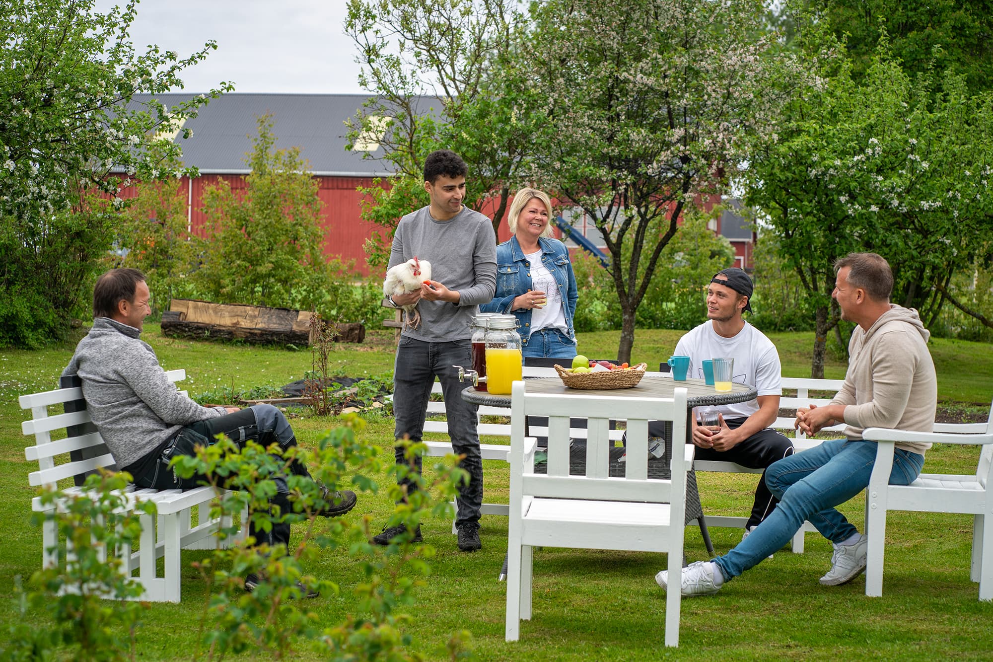 Fem personer samles rundt bord og benker i hage med frukt, drikke og en hvit høne. Smil og samtale i grønt landskap med rød bygning i bakgrunnen.