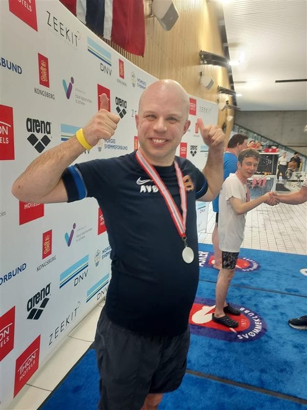 Anders, smilende med medalje og hevede never foran sponsorvegg etter svømmekonkurranse.