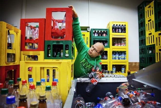 Anders i grønn KIWI hettegenser sorterer plastflasker i et pantesystem, omgitt av fargerike kasser og flasker.