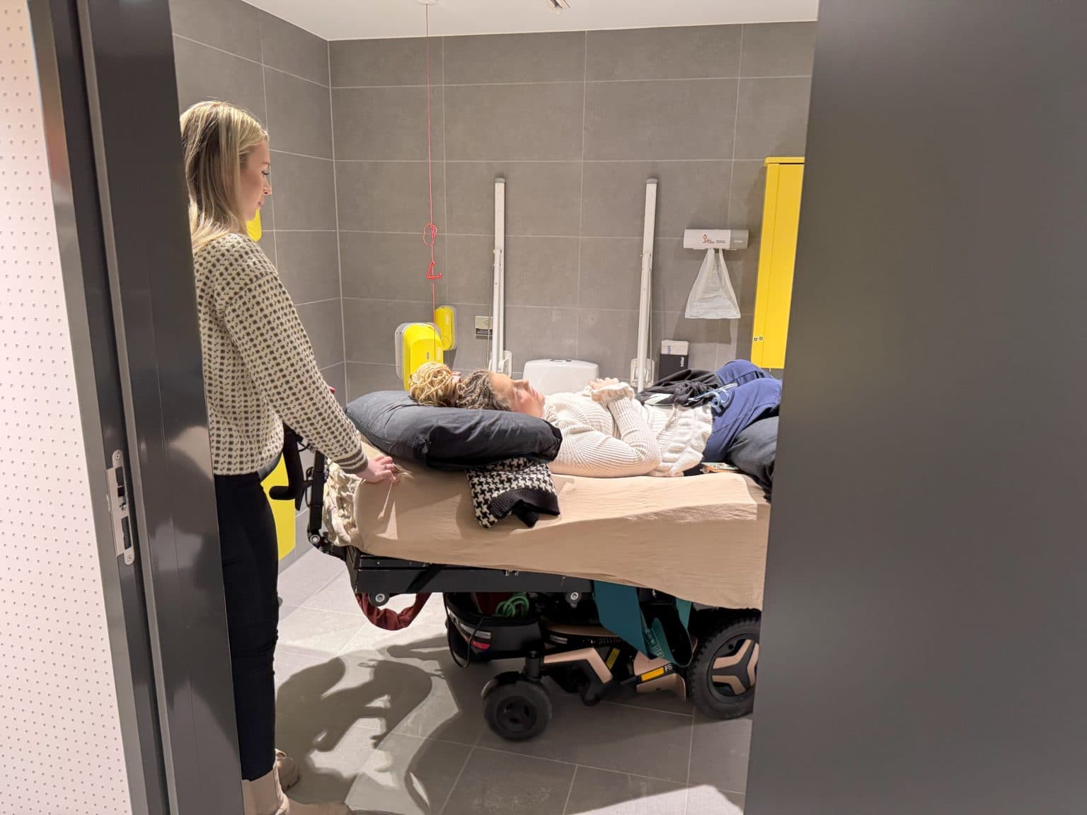 Person ligger på en mobil pleieseng i et moderne og tilgjengelig omsorgsrom. En annen person står ved siden av og gir støtte. Rommet har grå fliser, gult skap, vask, gripehåndtak og løftesystem med rød stropp.