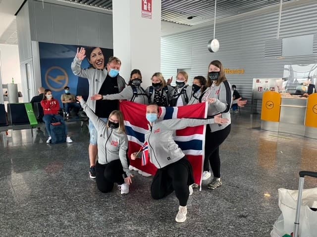 Ni personer poserer sammen i flyterminal, de fleste med grå jakker med røde og hvite logoer. To foran holder stort norsk flagg. Noen har munnbind. Bak sees Lufthansa-skranke og sitteområder.