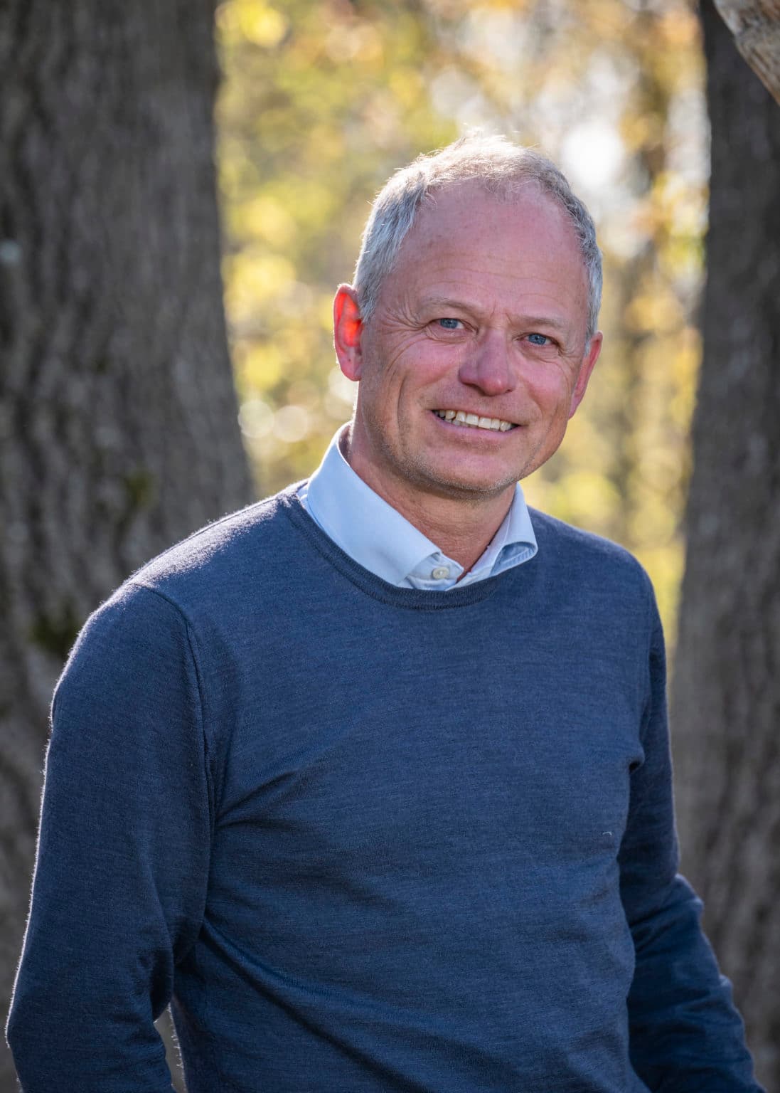 Kjetil i blå genser står foran trestammer i skogen, med sollys og høstfarger i bakgrunnen.