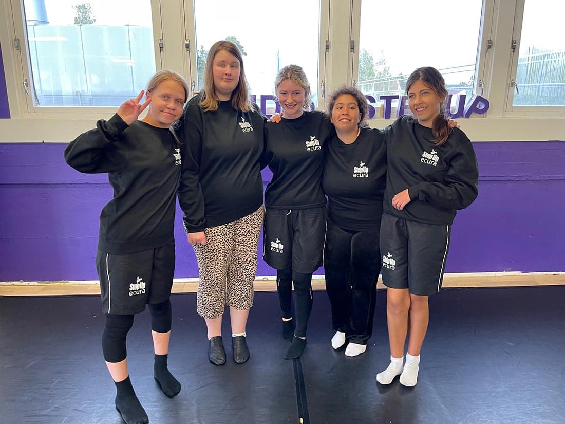 Fem personer står tett sammen foran lilla vegg med vinduer, alle i svarte gensere med logoen "Step Up ecura". Én gjør peace-tegn. Gulv og omgivelser tyder på treningsstudio.