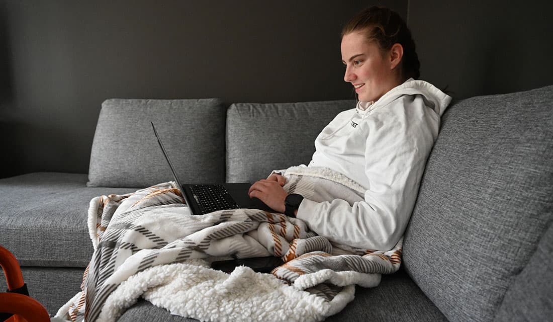 Person med hvit hettegenser sitter på grå sofa med mønstret pledd og svart laptop på fanget. Bakgrunnen er mørk og enkel.