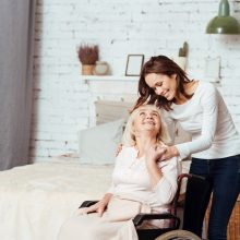 Positive young woman taking care of her grandmother in wheelchair Nice carer. Joyful delighted old woman sitting in wheelchair and lookign at her granddaughter who is takign care of her
